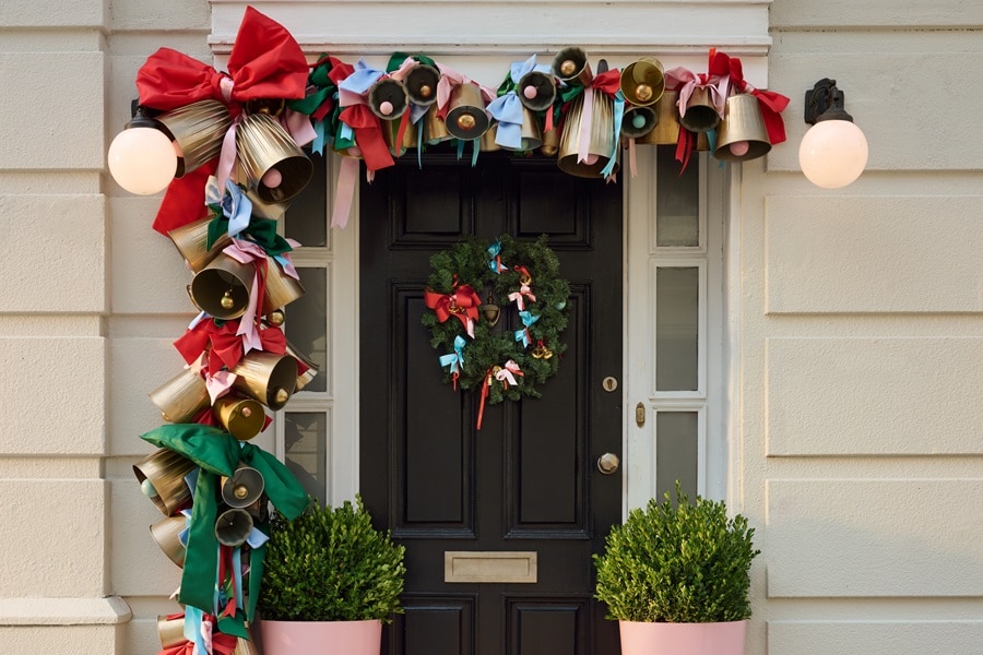 Porta d’ingresso con una ghirlanda di campanelle dorate e nastri rosso‑verdi appesa con i ganci Command Outdoor, e una corona su un gancio Command per ghirlande.
