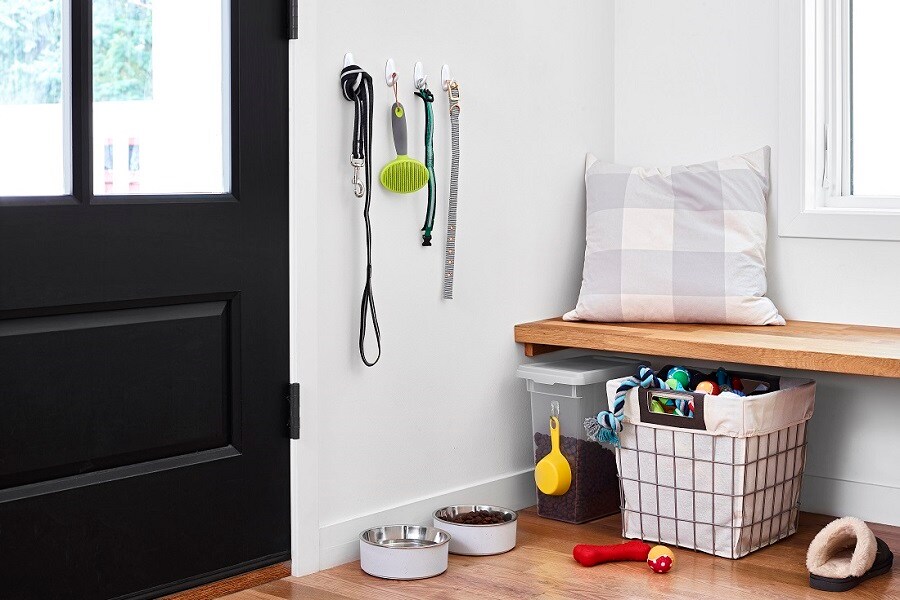Ingresso ordinato con ganci Command che tengono guinzaglio, spazzola e collare per cane. Sotto ci sono ciotole, un contenitore e un cesto per giocattoli. Una panca imbottita sopra.
